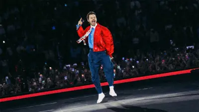 Harry Styles ma na sobie granatowe spodnie, jasnoniebieską koszulę z białym kołnierzykiem i krawatem w kwiatowy wzór oraz czerwoną kurtkę. Skacze na scenie, a za nim widać fanów obserwujących występ.