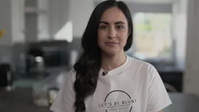 Leanne Lucas, who has long dark hair, smiles at the camera while wearing a white t-shirt with the logo 'Let's Be Blunt - Remove the Point'
