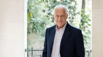 Antonio Fagundes, um homem de cabelos brancos, posa diante da câmera em um ambiente iluminado e cercado por vegetação. Ele veste blazer azul-marinho sobre uma camisa clara xadrez e aparece com expressão serena, em frente a uma varanda com fundo desfocado de plantas e árvores.
