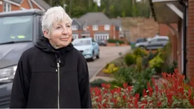 Irene Taylor looks out of the shot, has grey hair, and wearing a black hoodie. She stands in front of a flower bed and cars are behind her on the housing estate