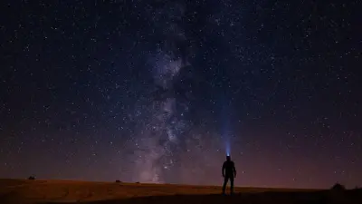 Uma pequena silhueta de uma pessoa usando uma lanterna de cabeça, olhando para um vasto céu noturno repleto de inúmeras estrelas, com a Via Láctea riscando o céu ao centro.