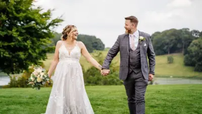 A newlywed couple hold hands and smile at each other against a backdrop of hills and trees. The groom is wearing a grey suit and the bride, who is holding some flowers in her other hand, is wearing a traditional white dress.