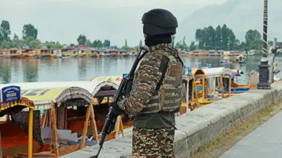 A paramilitary soldier carrying a gun standing by a river. There are houseboats in front of him