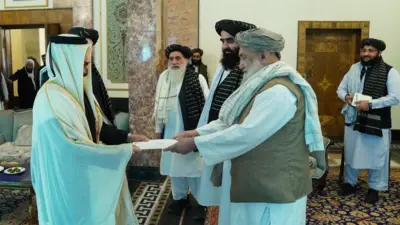 راست سفیر قطر که لباس مخصوص عربی سفید بر تن دارد و در مقابلش رئیس الوزرای حکومت طالبان که لباس سفید، واسکت روشن، لنگی نقرهای و دستمال سفید رنگ بر شانه دارد در حال گرفتن اعتمادنامه سفیر قطر است. آنطرف چند مقام دیگر طالبان ایستاده اند.