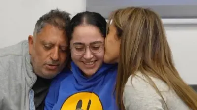  Liri Albag wearing a blue hoodie with a yellow smiley face sits in between her parents who are embracing and kissing her.