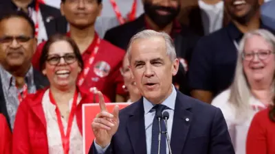 Canada's Prime Minister Mark Carney speaks at the Liberal Party election night headquarters. Behind him is a crowd of supporters. He is raising his hand and has a smile on his face. 