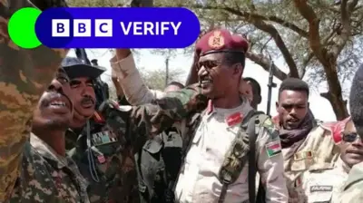 Former RSF commander Abu Keikal seen surrounded by troops. He holds a firearm aloft and is wearing a uniform and sunglasses. The men surrounding him are also wearing uniforms. 