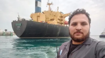 A selfie of a man in a grey overall taken in front of a vessel on a cloudy day