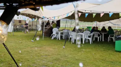 Marquee with white plastic tables and chairs underneath and people sheltering from the rain