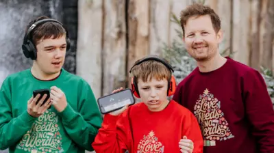 James com seus dois filhos Jude e Tommy. Os meninos estão com fones de ouvido e segurando um celular. Todos eles estão vestindo um suéter de Natal 