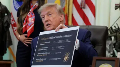Trump, sentado diante de mesa e com olhar sério, segura cartaz com novas recomendações sobre vacinação infantil.