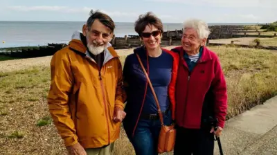 Colin, Christine and Beryl Bell are standing in front of a beach. Colin - an older, bearded man on the left of the three is smiling. He's wearing an orange raincoat. Christine, in the middle of the three, is wearing a blue coat and sunglasses. She has short brown hair. On the right, Beryl is an older woman in a red fleece coat. She is smiling at Christine and holding onto a walking stick.