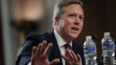 Дрісколл у темному діловому костюмі сидить за столом із мікрофоном, перед ним кілька пластикових пляшок з водою. Руки підняті з розкритими долонями, ніби в жесті пояснення або зупинки.