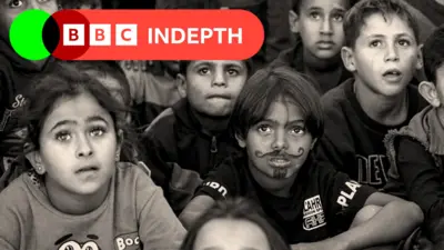 An image in black and white of young children sitting on the ground and looking up, one of them has a beard and moustache drawn on his face in pen