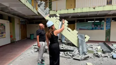 Claudia Arenas e um homem estão no pátio de uma escola da qual parte do telhado se desprendeu.