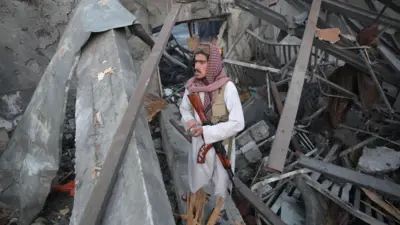 A Taliban security guard stands at the scene of alleged Pakistani air strikes in Kabul, Afghanistan, on 17 March 2026. He holds a smartphone in his hands, and a gun is slung over his shoulders.