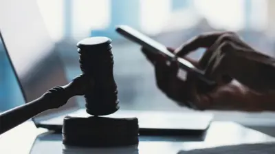 Mão mexe em celular em frente a computador, com martelo da Justiça em primeiro plano