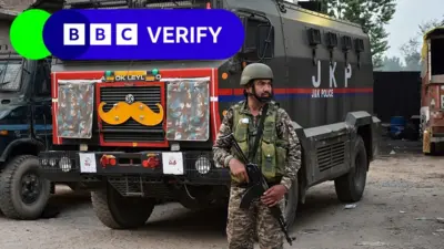 An Indian soldier stands guard. He is armed with a rifle and is wearing army fatigues. In the background is a large truck.