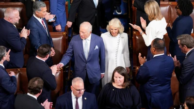 King Charles wearing a blue pinstripe suite, white shirt and pale blue tie walks down an aisle shaking hands with people after delivering his sppech. He is viewed from above and Queen Camilla is next to him wearing white