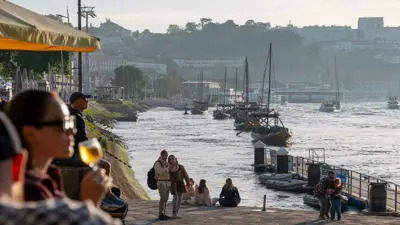 Apus de soare la Porto, în nordul Portugaliei. Oameni stau pe malul fluviului Douro. Una dintre femei, în prim-plan, în stânga, ține în mână un pahar cu vin. Alții se plimbă pe faleză și fac fotografii.
