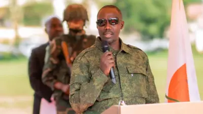 Sultani Makenga, yambaye imyenda y'amabara y'icyatsi kibisi y'igisirikare cya Congo n'amataratara y'umukara afite indangururamajwi mu ntoki arimo kuvuga, inyuma ye hari umusirikare umurinze.