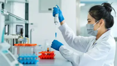 Kép egy női kutatóról egy laboratóriumban. Sötét haja magas kontyba van fogva, fehér köpenyt, gumikesztyűt és arcmaszkot visel. Egy tűvel folyadékot fecskendez kémcsövekbe.