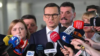 Mężczyzna z ciemnymi, krótkimi włosami ,w czarnych okularach, białej koszuli i w niebieskiej marynarce stoi w otoczeniu pięciu osób, przed nimi znajdują się mikrofony prasy