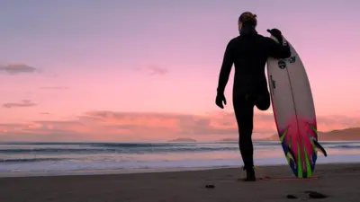 Surfista veste uma roupa de neoprene preta de corpo inteiro e luvas. Ele tem apenas uma perna e está de pé na praia, diante de um pôr do sol em tons de rosa. De costas para a câmera, olha para o mar e para a imensidão do céu. Segura uma prancha de surfe branca, rosa-choque e verde, com a ponta voltada para o céu e a outra extremidade apoiada na areia
