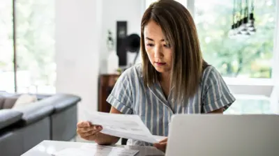 A woman looks at a paper bill while using a laptop at home. She's wearing a blue stripey top and her home is bright and modern.