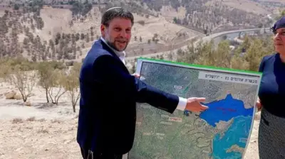 : بتسالل اسموتریچ، وزیر دارایی اسرائیل، نقشه بزرگی را در دست گرفته رو به دوربین نگاه می‌کند و با دست به نقطه‌ای از نقشه اشاره می‌کند. زنی هم در کنار نقشه ایستاده است
