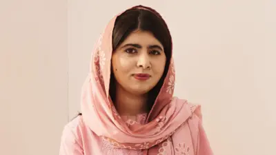 Malala Yousafzai, posing for the camera while wearing traditional clothing in a pale pink colour with matching headscarf.  