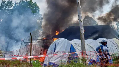Fire and smoke at the of burnt tents on the grounds of Rwampara General Hospital. A police officer with a gun can be seen to the right of the shot in front of other makeshift tents.