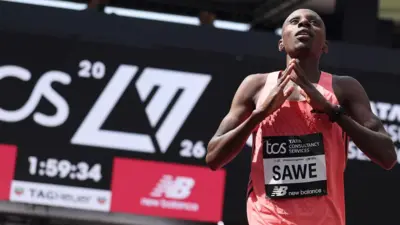 ランニングウェア姿で胸にSAWEと書かれたゼッケンをつけたサウェ選手が、胸の前で両手の指先を合わせている。背後には1:59:34のタイム表示がぼんやり見える