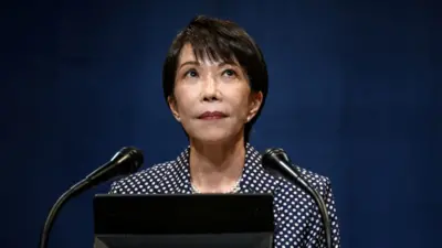 黒地に白い水玉模様のジャケットを着た高市氏が、2本のマイクに向かって立っている