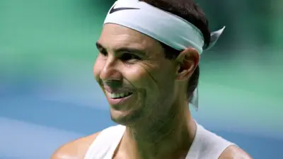 Tennis player Rafael Nadal smiles broadly during a training session. He has short dark hair, tied back with a sweatband, and is dressed in a white, sleeveless top.