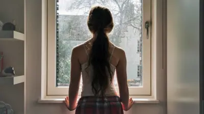 Stock image of a girl with long hair tied in a ponytail stands next to a window and looks outside, wearing a vest and checked pyjama bottoms.