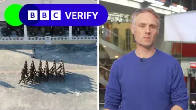 A composite image split in half showing soldiers on the left, and BBC Verify reporter Richard Irvine-Brown on the right. On the left, a group of around 30 Syrian soldiers in dark green uniform are walking in a tight square formation through a large courtyard. The afternoon sun casts long shadows. The group are being watched by several senior ranking officers who sit under a balcony. The BBC Verify logo is in the top left. On the right hand side of the composite, Richard wears a dark blue jumper and has a serious expression. He is standing in the BBC London newsroom. Behind him is a spiral staircase and several television screens.
