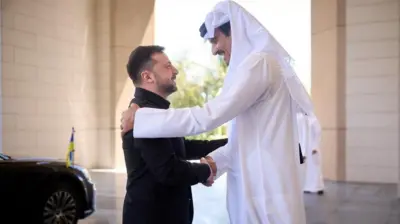 Ліворуч президент Зеленський у чорному костюмі, напроти тисне йому руку та усміхається - емір Катару Тамім бін Хамад Аль Тані у білому мусульманському вбранні та білій куфії. 