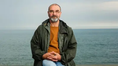 Andrew Malkinson yicaye afatanirije amaboko ku bibero. Yambaye ipantaro ya jeans, umupira w'ibara rigomba gusa n'umucungwe uhiye, n'ikoti isa n'icatsi kibisi, kandi ariko araba kamera adatwenga. Inyuma yiwe hari ikiyaga.