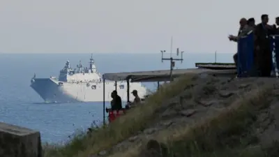 Kıyıdaki bir yükseltide insan silüetleri görünüyor ileride denizde bir donanma gemisi var.