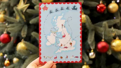 A map of the United Kingdom with a Christmas-themed design, showing a trail of Santa’s sleigh and various holiday icons such as reindeer, robins, and other animals, held in front of a decorated Christmas tree with ornaments