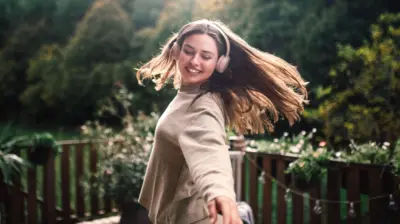 yeşillikler içinde kulaklıkla dans eden genç bir kadın. Saçları uzun, düz ve kumral. Üzerinde krem rengi uzun kollu bir üst var.