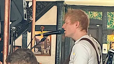 Singer Ed Sheeran at the microphone wearing a white T-shirt singing in a pub