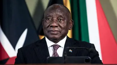 South Africa President, Cyril Ramaphosa wear black suit, brown neck-tie wey get designs and white inner shirt. E sand for podium as e look inside di camera.