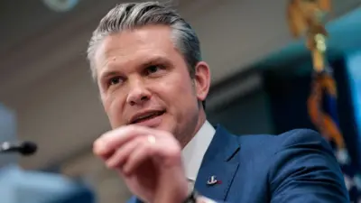 Foto de Pete Hegseth dos ombros para cima, no púlpito da sala de imprensa do Pentágono. Ele veste um terno azul-petróleo bem ajustado sobre camisa branca. Sua mão esquerda está erguida, com o polegar e o indicador unidos, como se enfatizasse um ponto