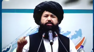 وزیر تحصیلات عالی حکومت طالبان با لنگی و واسکت سیاه و پیراهن سفید پشت مایک در حال صحبت کردن است.