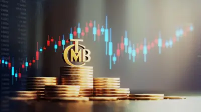 madeni paralar üst üste dizilmiş üzerinde TCMB amblemi var hepsi altın rengi ve arkada borsa işlemleri var mavi ve kırmızı bir grafik.