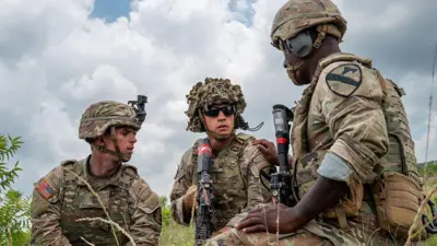 Three US soldiers inside one field, dem wear helmets and dem dey hold weapons and tactical gear