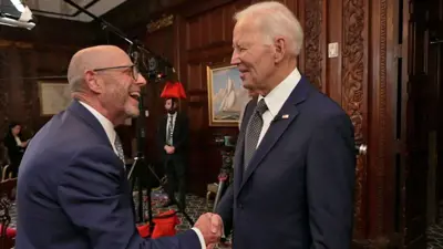 Joe Biden wears a dark blue suit and shakes hands with BBC journalist Nick Robinson