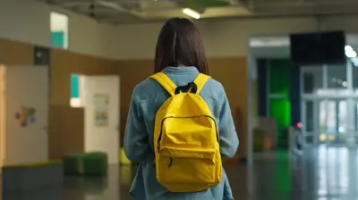 Дівчина із жовтим наплічником на спині стоїть у порожньому холі школи.